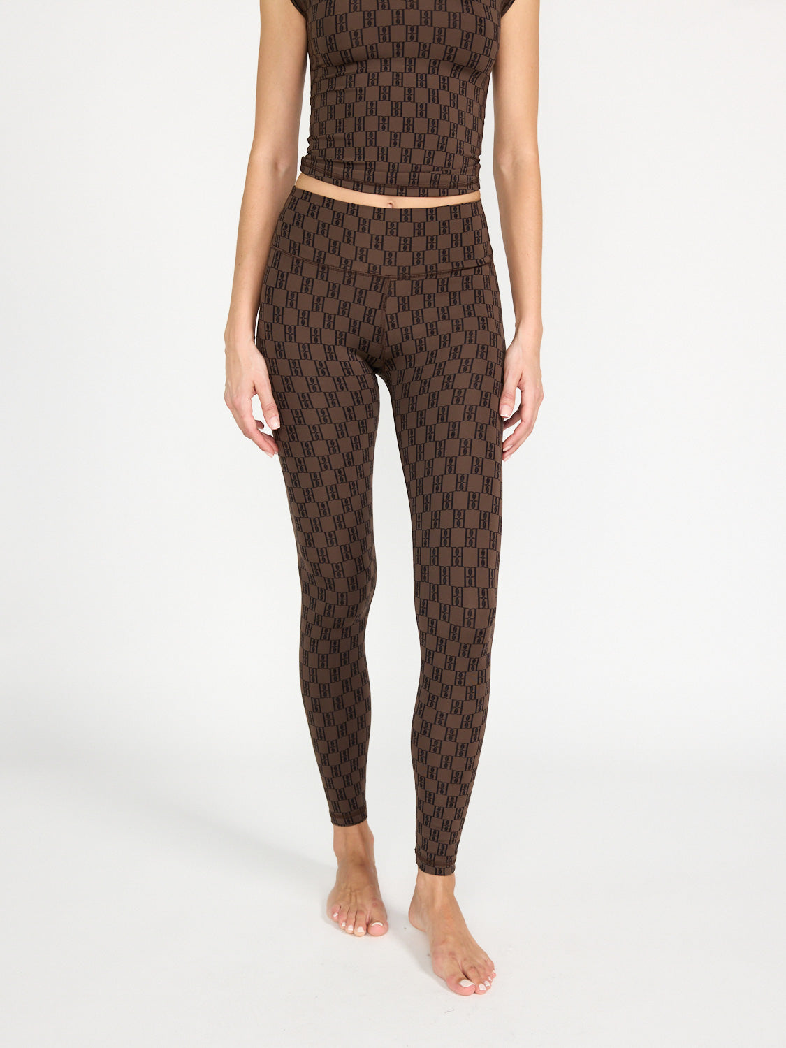 Eine Frau steht barfuß vor einem schlichten Hintergrund und trägt by malene birger Polene - Leggings mit Brand-Details und ein passendes Crop-Top mit schwarzem geometrischen Muster. Ihre Arme liegen entspannt an den Seiten, und das Bild ist an den Schultern abgeschnitten.