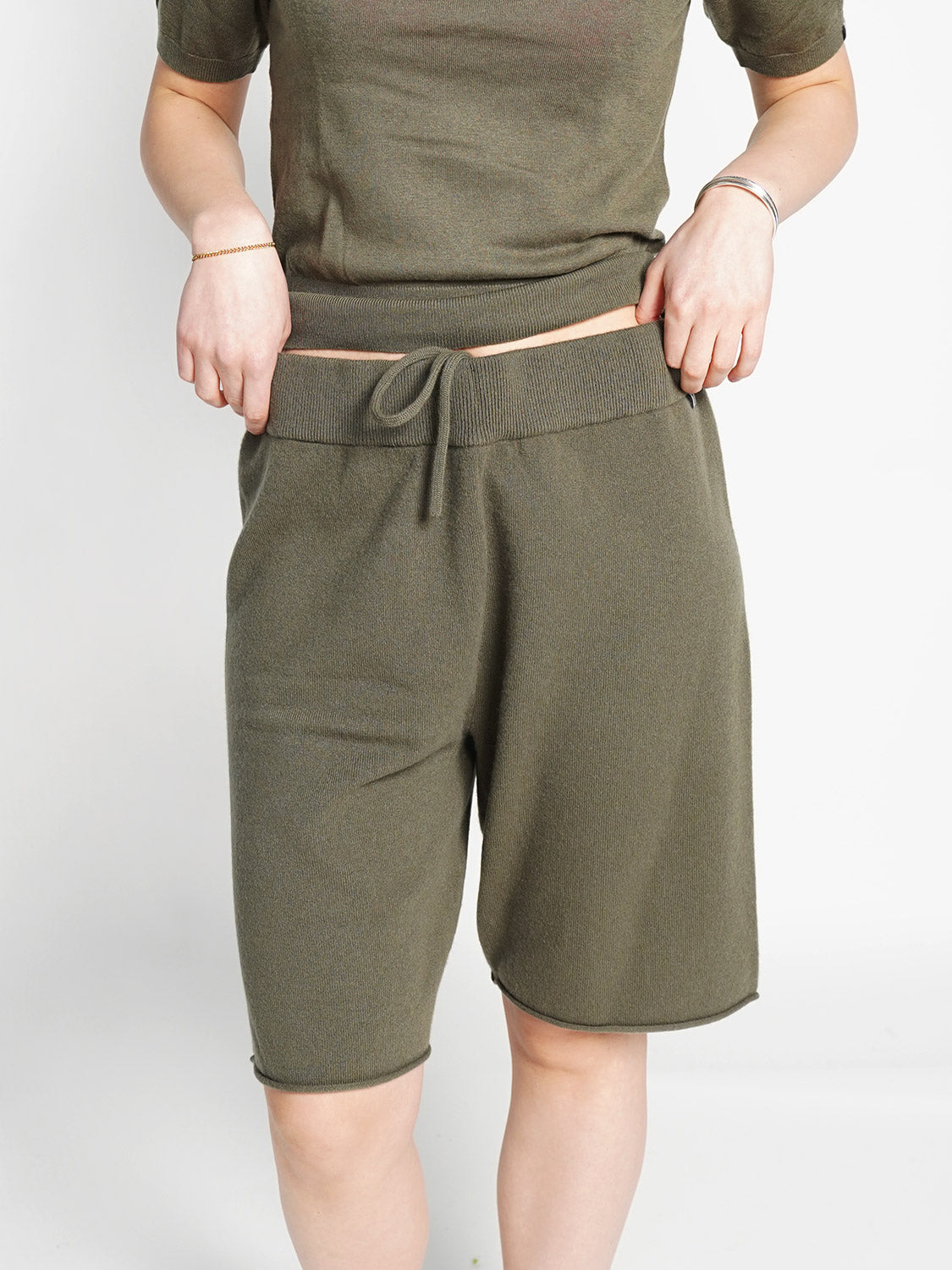 Eine Person modelliert die Extreme Cashmere Laufen - Shorts aus Cashmere, ein olivgrünes, knielanges Modell mit Kordelzug in der Taille, gepaart mit einem passenden kurzärmeligen Oberteil. Das Gesicht ist auf dem einfarbig weißen Hintergrund nicht zu erkennen.